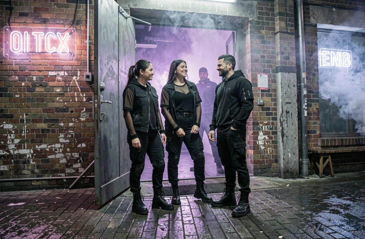 Groupe d'amis en tenue techno streetwear devant l'entrée d'un club industriel la nuit avec reflets néons sur pavés mouillés
